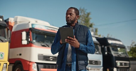 Attractive African American male driver using his work tablet device while standing before trucks....
