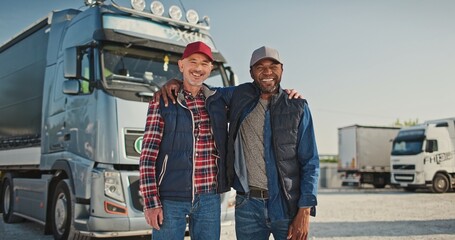 Portrait of happy professional workers, truck drivers. Two multicultural male colleagues looking at camera and smiling. Confident lorry drivers with vehicle in parking lot. Truck car travels © VAKSMANV