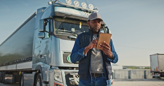 African American professional truck driver setting up navigation for destination. Checking his route on tablet computer and standing by long vehicle. Transportation service.