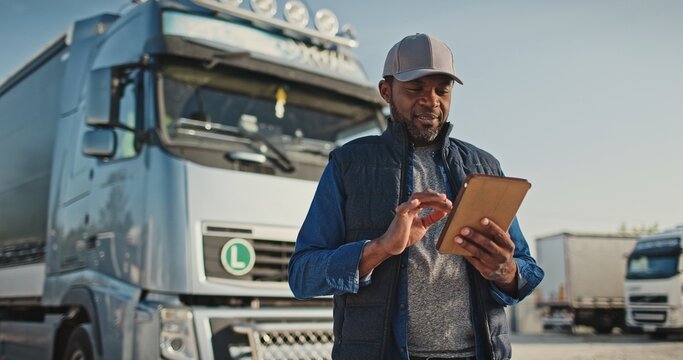 African American professional truck driver setting up navigation for destination. Checking his route on tablet computer and standing by long vehicle. Transportation service.