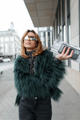 Glamorous beautiful stylish woman model with fashion glasses in a shaggy jacket with a magazine and smartphone walks in the city