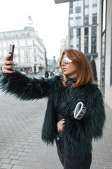 Fashion beautiful glamorous girl in stylish clothes takes a selfie on a smartphone and walks in the city