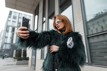Fashion beautiful stylish girl model with cool sunglasses in a fashionable outfit with a shaggy jacket holds a magazine and takes a selfie photo on a smartphone