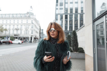 stylish urban beautiful fashionable girl with glasses in a fashion shaggy jacket with a magazine and a smartphone walks in the city