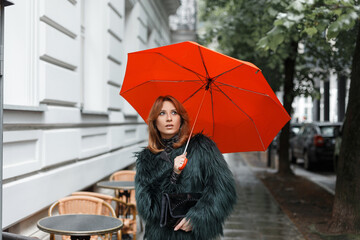 Beautiful fashionable woman model in fashion clothes with handbag with orange umbrella walking on a rainy day in the city