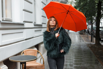 Fashionable beautiful girl model in stylish clothes with a jacket with a bright orange umbrella walks in the city on a rainy day