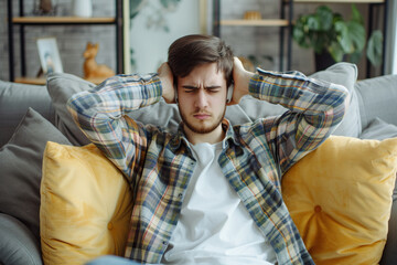 Dissatisfied and angry young man sitting on sofa at home and covering ears from excessive noise