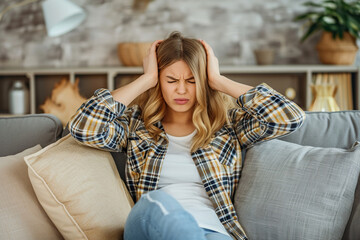 Obraz premium Dissatisfied and angry young woman sitting on sofa at home and covering ears from excessive noise