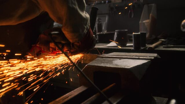 A blacksmith in the forge uses a grinder to shape a piece of metal into the form of a new product. He wears a protective mask to protect his face from flying sparks due to sawing