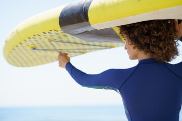 Positive woman in curly hair lifting yellow paddleboard - 796517130