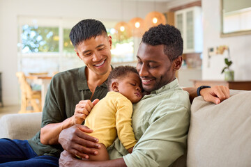 Same Sex Male Couple Or Friends Cuddling Sleeping Baby Daughter On Sofa At Home Together