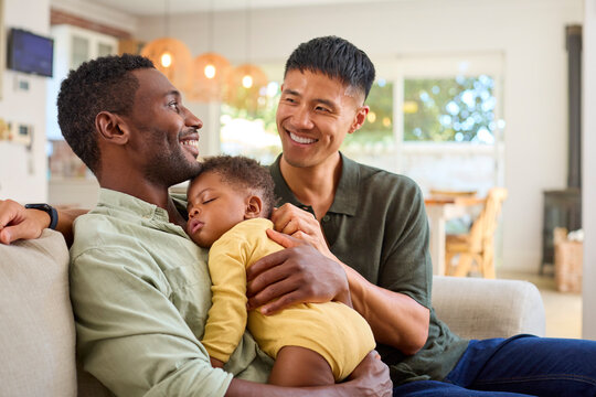 Same Sex Male Couple Or Friends Cuddling Sleeping Baby Daughter On Sofa At Home Together