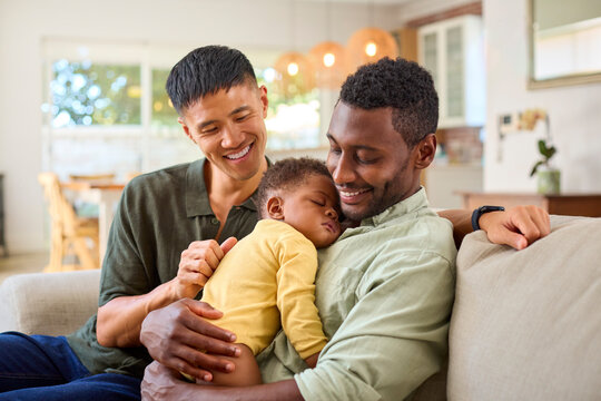 Same Sex Male Couple Or Friends Cuddling Sleeping Baby Daughter On Sofa At Home Together