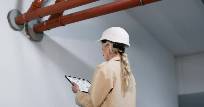 Female engineer in helmet holds tablet with plan examining fire protection system. Woman worker engages in building code inspection procedure