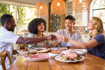 Group Of Multi-Racial Friends Sitting Around Table Enjoying Meal At Home Making Cheers With Water