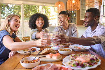 Group Of Multi-Racial Friends Sitting Around Table Enjoying Meal At Home Making Cheers With Water