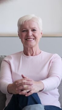 Portrait of a smiling Caucasian mature woman sitting on sofa at home and looking at camera. Optimistic and cheerful people. Vertical video.