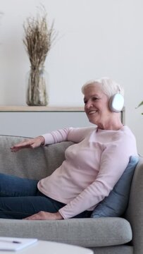 Mature happy Caucasian woman listening music in wireless headphones and relaxing on sofa at home. Recreation, pleasure, refreshment concept. Vertical video.