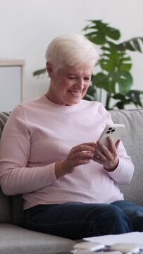 Mature Caucasian happy woman saying wow and showing thumbs up while using cellphone on sofa at home. Messaging online in social media. Vertical video.