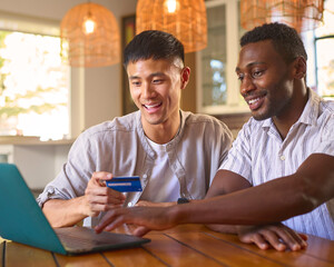 Same Sex Male Couple At Home With Laptop And Credit Card Booking Holiday Or Shopping Online