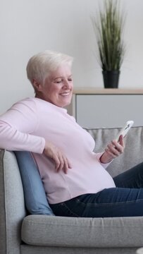 Senior Caucasian woman browsing internet with cellphone portrait and looking at camera on sofa at home. Resting, relaxation. Vertical video.