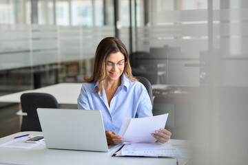 Happy busy mature business woman working in office checking legal account invoice. Businesswoman financial advisor consultant reading document, checking tax paper using laptop. Authentic shot.