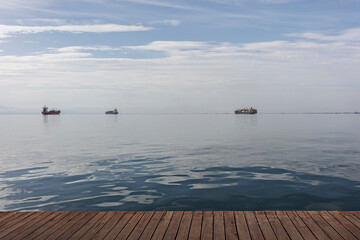 A wooden pier in Thessaloniki, Greece foregrounds a serene ocean vista where distant cargo ships...