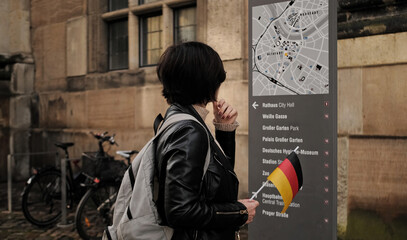 Attractive Female Tourist Walks With Germany Flag In Historical Center Of Dresden, Checking Touristic Route On City Map