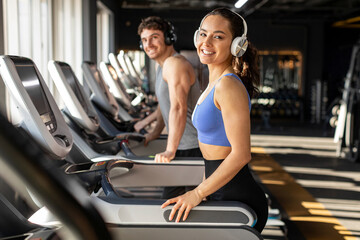 Cheerful woman in sportwear and headphones exercise running treadmill in fitness gym health club, smiling at camera. Calories burn during cardio training