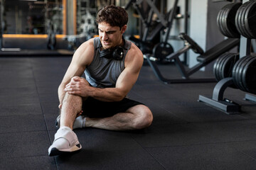 Sport trauma. Man touching his hurt knee at gym, sitting on floor and massaging sore injured leg in...