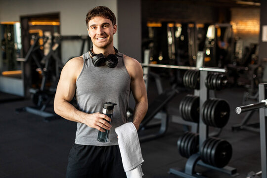 Sporty man holding water bottle and towel after sport workout at modern gym, guy relaxing after training in fitness club, smiling at camera