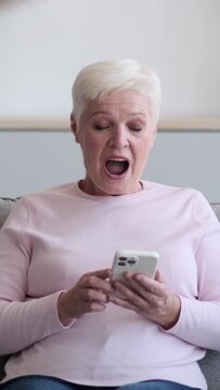Surprised and cheerful Caucasian elderly woman using phone saying Wow and showing thumbs up at home. Spending leisure time with online entertainment. Vertical video.
