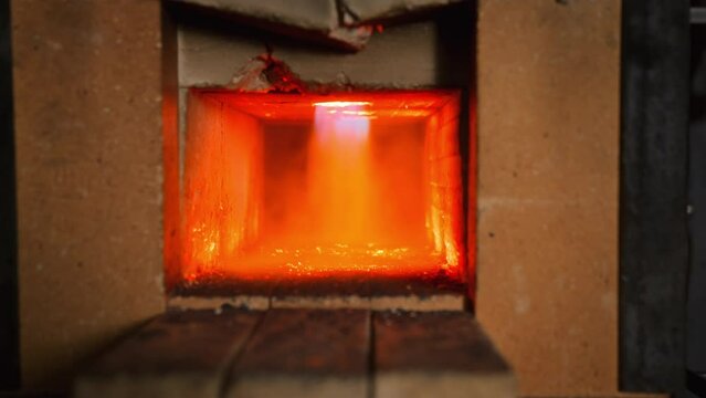 A close up of a stone furnace in a forge, with a red flame flaring up. A metal product is being pushed inside to be heated and melted.