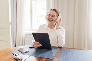 Joyful young professional wearing headphones and holding a tablet, engaging in a video call at her...