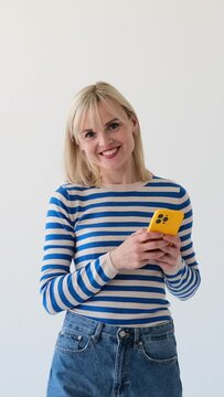 Portrait of a happy and smiling Caucasian woman with phone browsing internet and looking at camera on white background Vertical video.