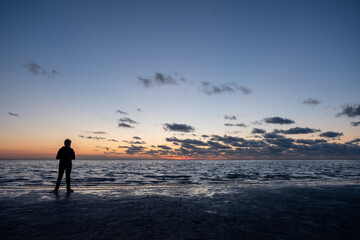 A solitary silhouette stands on the expansive seashore, gazing at the sunset where the sky meets the ocean. The horizon is painted with warm hues fading into the cool blues of twilight, while