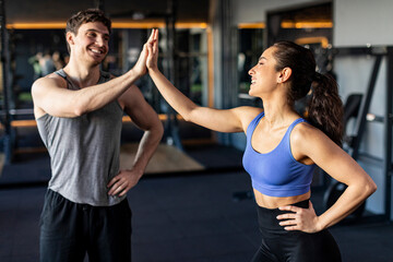 Athletic fitness lady and coach man give high five during training in dark gym interior, smiling at...