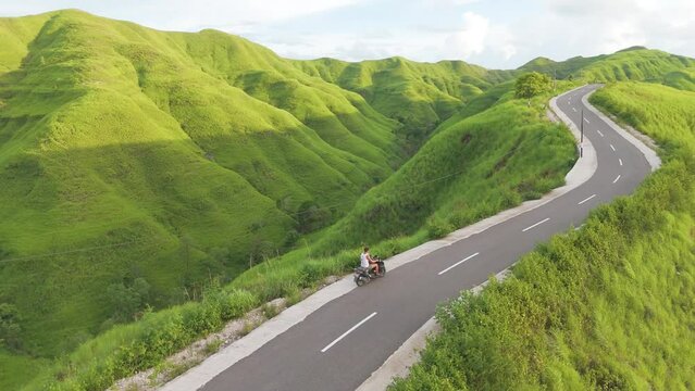 Sumba, Indonesia. A man is riding a motorcycle down a road with a beautiful view of the mountains in the background.