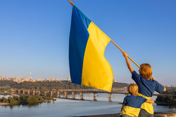woman and child with yellow-blue flag stand high on the roof of a house against of sky and Dnieper...