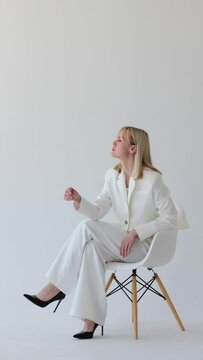 Pensive Caucasian middle aged businesswoman in doubt, sitting on chair and contemplating, planning strategy or answering on question on white background. Vertical video.