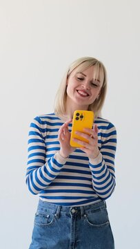 Cheerful Caucasian woman using phone for texting in online social media and laughing, spending leisure time on white background. Vertical video.