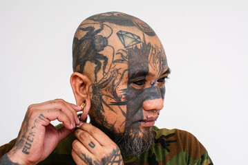 A bald asian man attaches a large captive bead ring to his flesh tunnel. Example of extensive facial tattoos and body piercings or modifications.