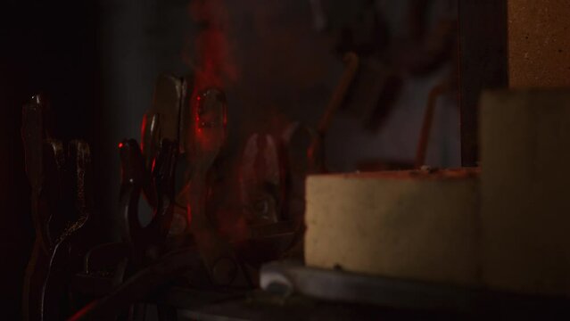 A close up of a stone furnace in a forge, with a red flame flaring up. A metal product is being pushed inside to be heated and melted.