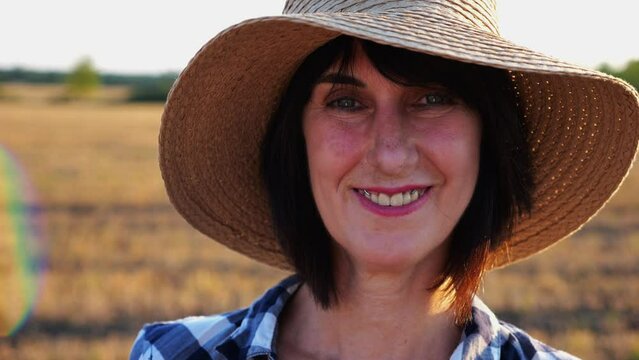 Happy smiling female farmer looks into camera standing at wheat field. Portrait of adult beautiful agronomist with straw hat with barley meadow at background. Agricultural business concept. Close up