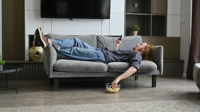 A young red-haired guy is lying on the sofa and eating corn flakes, scrolling through social networks on his phone