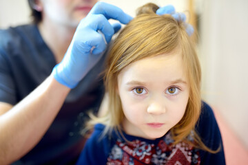 Close-up portrait of little girl during appointment of dermatologist in modern clinic. Doctor examines child hair and scalp for lice and nits. Pediculosis is common disease in kids groups
