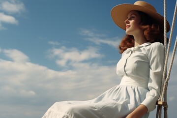 Stylish young woman in an elegant vintage dress and fashionable straw hat sitting gracefully on a wooden swing and gazing contemplatively into the distance under the serene blue sky on a sunny day