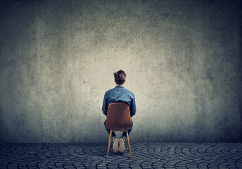 A man sits on a chair and looks at an empty concrete wall - obrazy, fototapety, plakaty