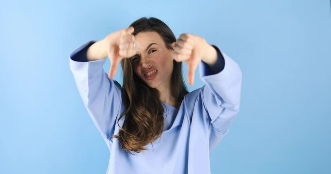 Displeased brunette young woman posing isolated on blue background studio. Woman in blue long sleeves top looking camera with finger gesture showing thumb down, tongue out.