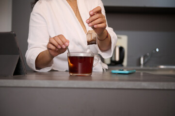 Close-up woman in white bathrobe, sweetening his tea, standing at kitchen table with open digital...